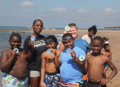 Summer camp students from the YWCA in New Britain, and Taraea Ellis, second from left, school-aged teacher at the YWCA, hold up stickers they received while visiting Lighthouse Point Park from the “Don’t Trash Long Island Sound – Break the Single Use Plastic Habit” campaign kickoff. Judy Benson / Connecticut Sea Grant